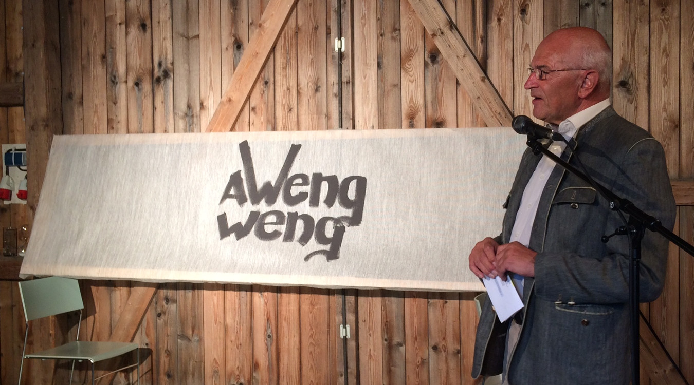 Bezirkstagspräsident Dr. Günther Denzler steht am Mikrofon vor einem Banner auf dem die Redewendung a weng weng zu sehen ist.