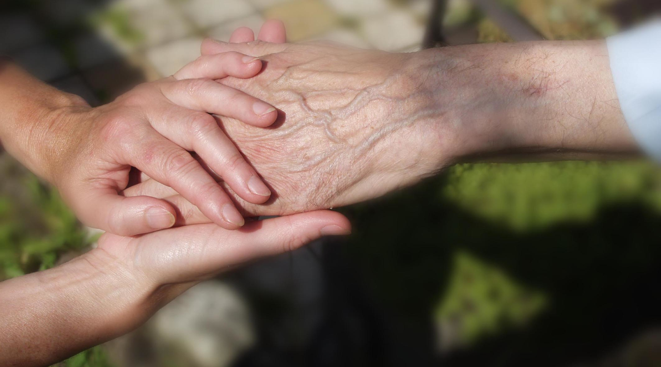 Zu sehen sind zwei Hände. Die Hand einer älteren Person wird von zwei Händen umschlossen. 