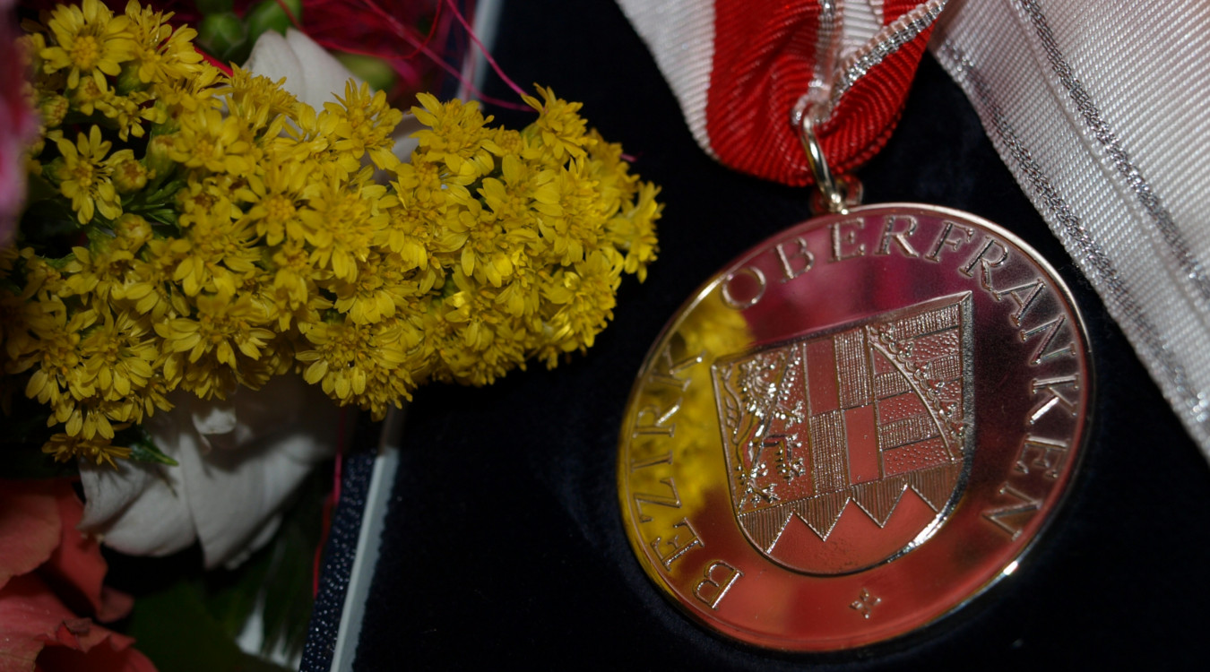 Das Bild zeigt die Ehrenmedaille des Bezirks Oberfranken in Silber. Die Medaille trägt das Wappen des Bezirks und hängt an einem rot-weißen Band.