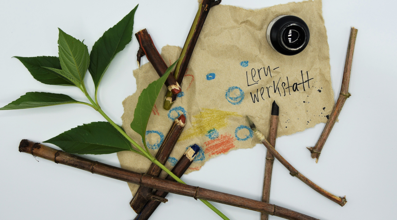 Äste, Wachsmalstifte, eine Pflanzen und ein Papier mit blauen Kreisen und dem Wort "Lernwerkstatt" liegen auf einem weißen Untergrund.