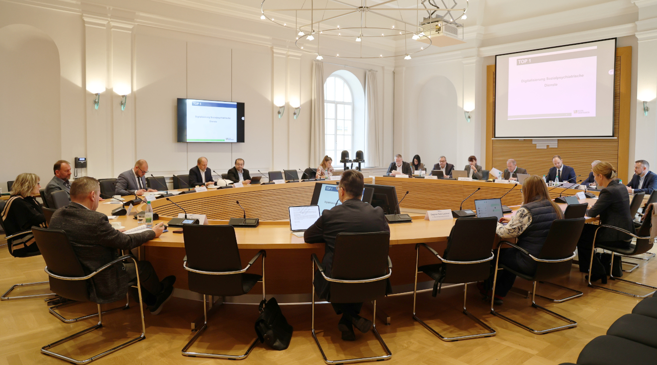 In einem großen Saal sitzen an einem runden Konferenztisch Menschen. Vor sich haben Sie Unterlagen und Mikrofone auf dem Tisch.
