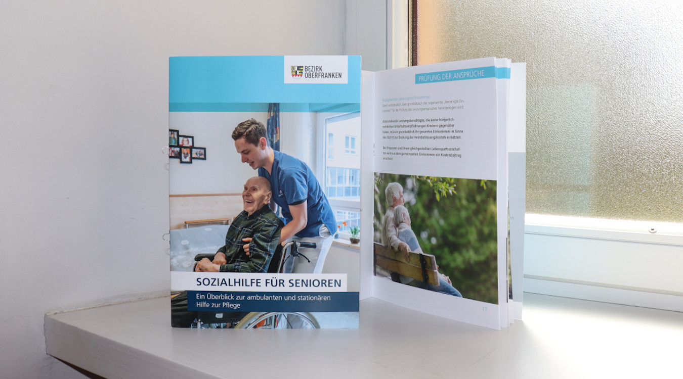 Das Bild zeigt das Deckblatt der Infobroschüre "Sozialhilfe für Senioren". Dahinter sieht man eine aufgeschlagene Seite. Die zwei Exemplare der Broschüre stehen auf einem weißen Fensterbrett.