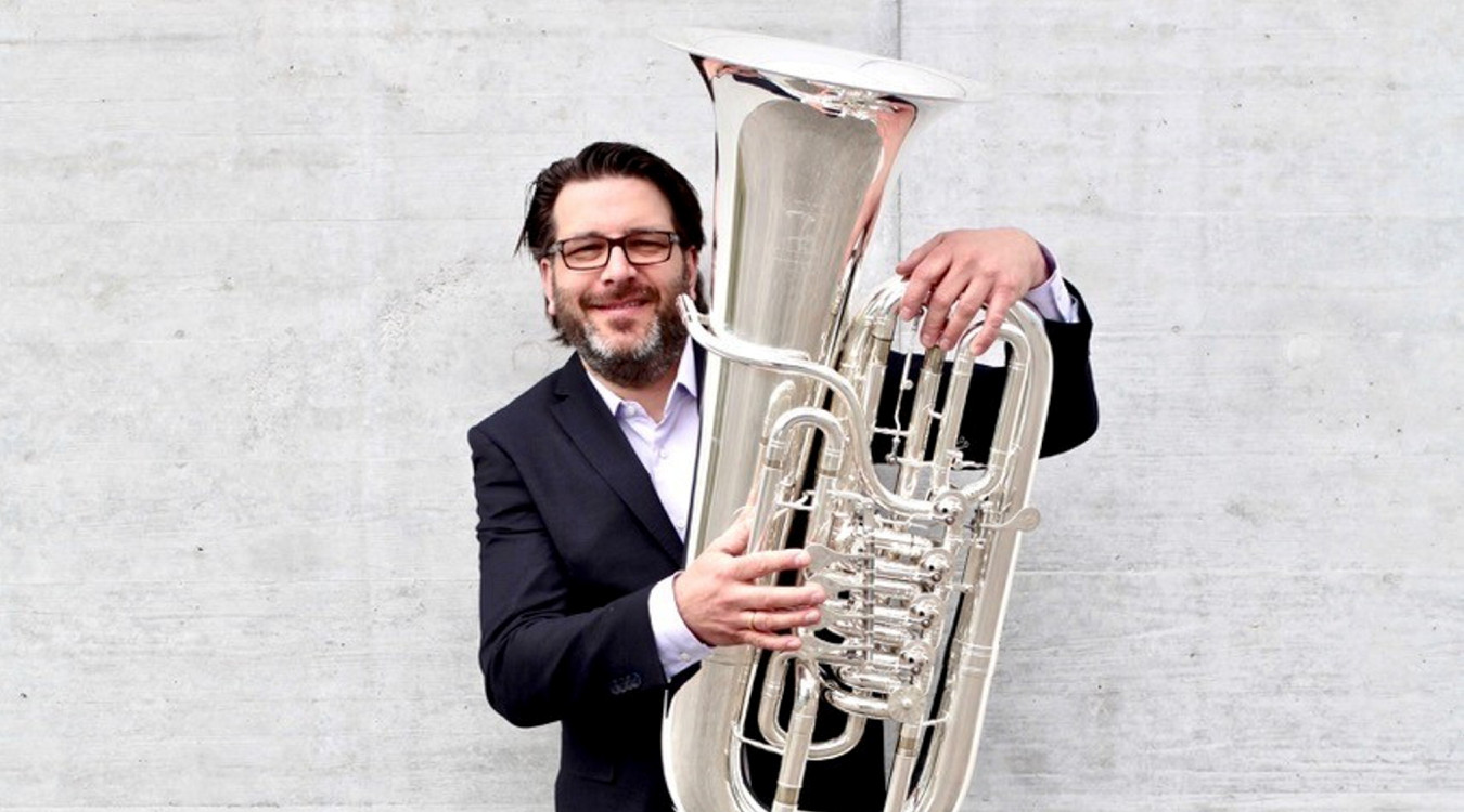 Ein Herr im Anzug steht mit angewinkeltem Bein an einer Mauer und blickt fröhlich in die Kamera. In den Händen hält er eine Tuba.