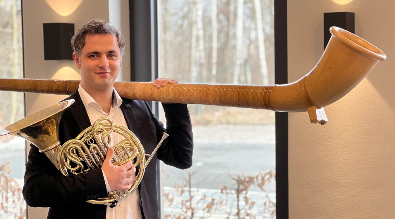 Ein Mann im Anzug hält ein Blasinstrument über der Schulter und ein Waldhorn in der Hand.