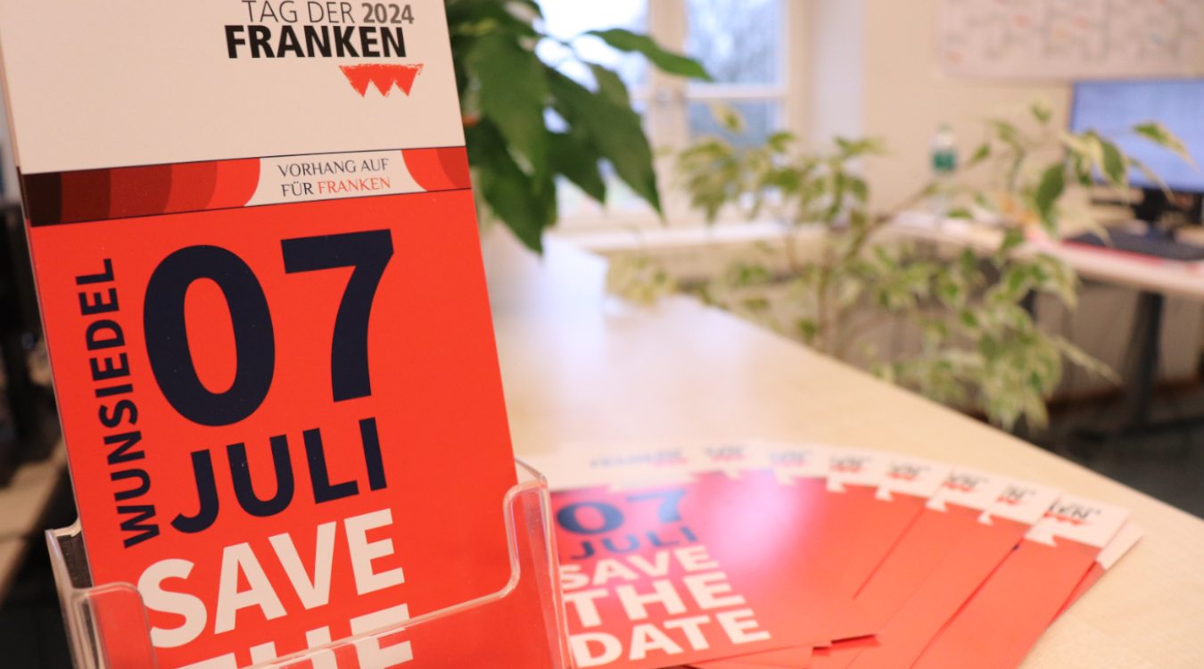 Save the Date: 7. Juli 2024 Rote Flyer vom Tag der Franken stehen auf einem Regal