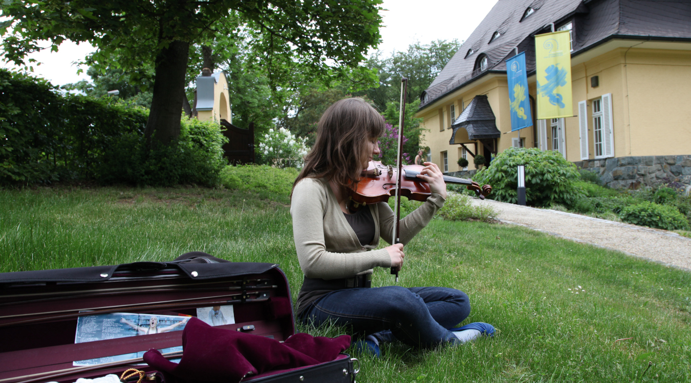 Eine junge Frau sitzt auf einer Wiese vor einem Haus und spielt Geige