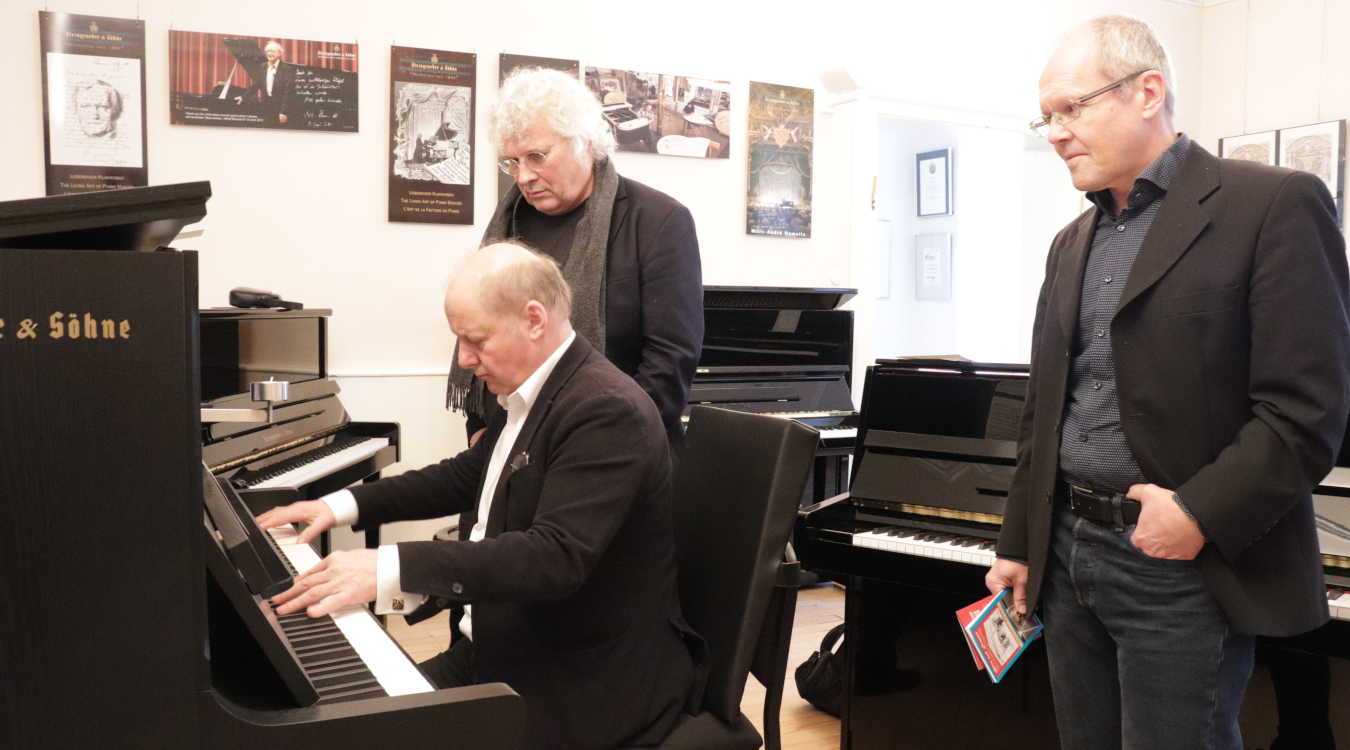 Zu sehen ist Prof. Wolfgang Manz am Klavier, dahinter stehen künstlerischer Leiter Prof. Christoph Adt und Verwaltungsleiter Dr. Ulrich Wirz.