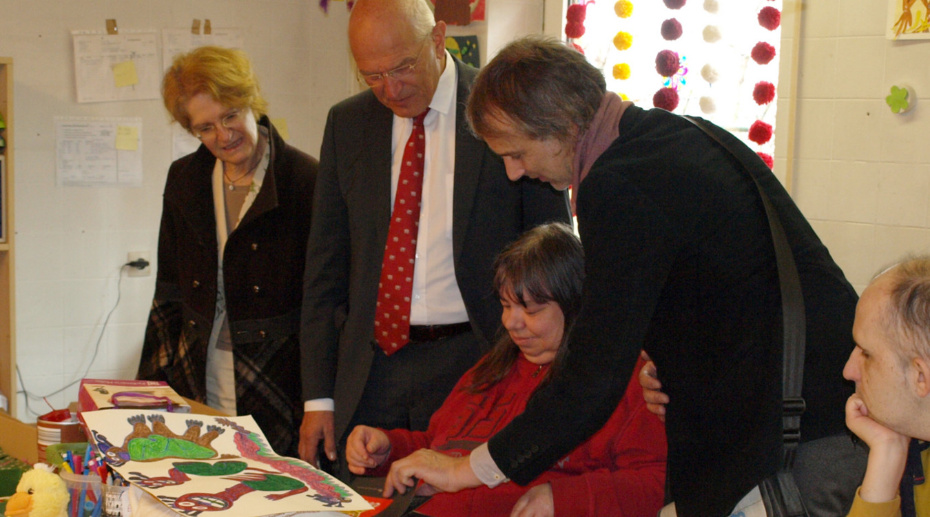 Bezirkstagspräsident Dr. Günther Denzler schaut sich in einer Einrichtung für Menschen mit Behinderung die Arbeit einer Frau an. Sie malt und gestaltet Kalender an einem großen Basteltisch.