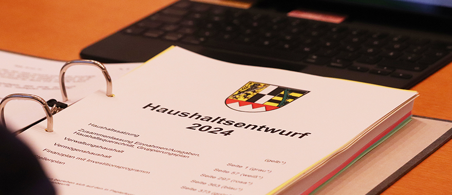 Auf einem Schreibtisch liegt eine mehrere hundert Seiten starkes Buch mit der Aufschrift Haushalt 2021 und dem Wappen des Bezirks Oberfranken. Im Hintergrund liegt ein Taschenrechner.