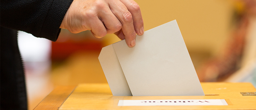 Eine Hand wirft einen Umschlag in eine Wahlurne.