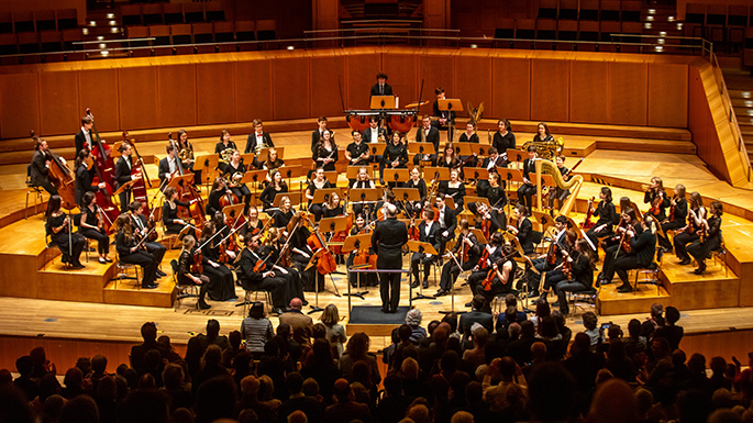 Ein Konzertsaal ist mit einem Orchester gefüllt. Man sieht das Publikum von hinten.