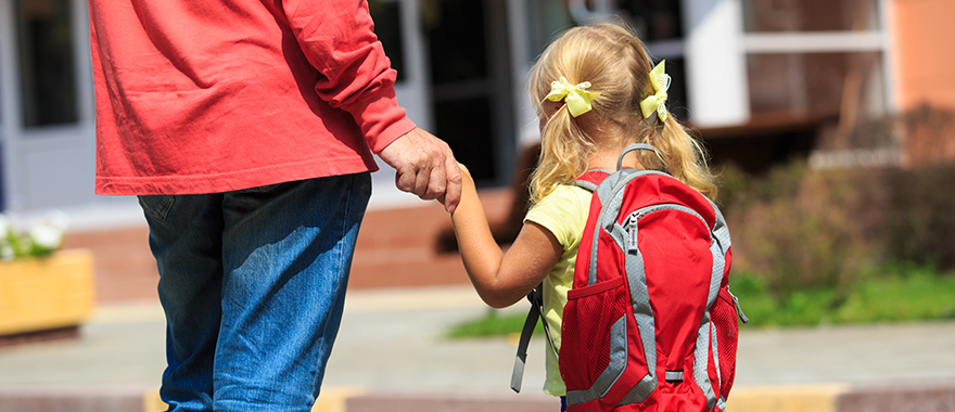 Ein Vater geht mit seiner Tochter, die einen Rucksack trägt, auf ein Schulgebäude zu.