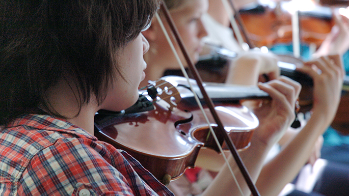 Ein Junge spielt auf einer Geige, im Hintergrund sitzen weitere Geigenspieler 