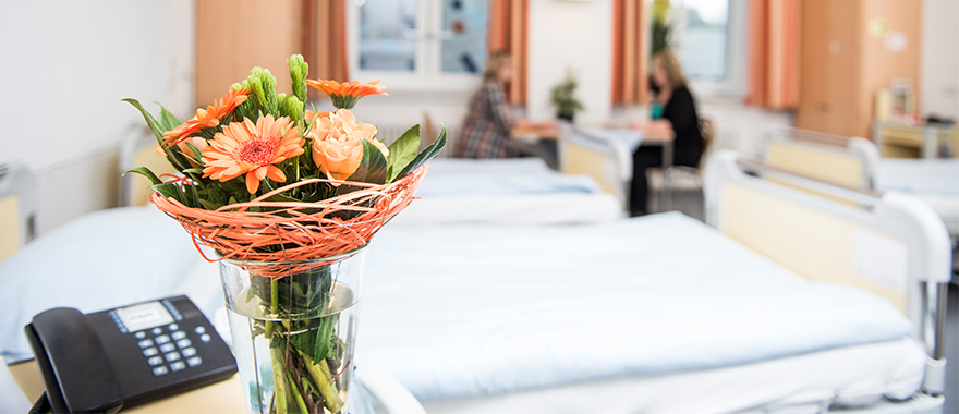 Das Bild zeigt ein helles Krankenzimmer des Bezirkskrankenhauses Bayreuth. Auf dem Nachtisch im Vordergrund steht ein Strauß frischer oranger Blumen und ein Telefon. Im Hintergrund sind unscharf zwei Menschen zu erkennen. 