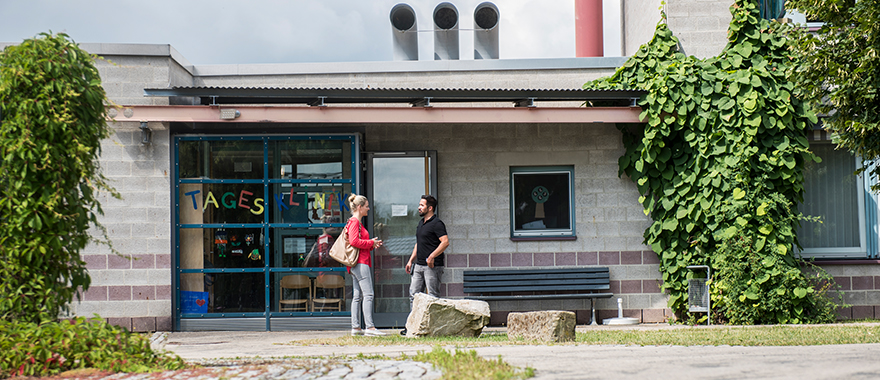 Das Bild zeigt das Gebäuede der Tagesklinik für Kinder- und Jugendpsychiatrie in Bayreuth von außen. Vor dem Eingang stehen zwei Personen und unterhalten sich. 