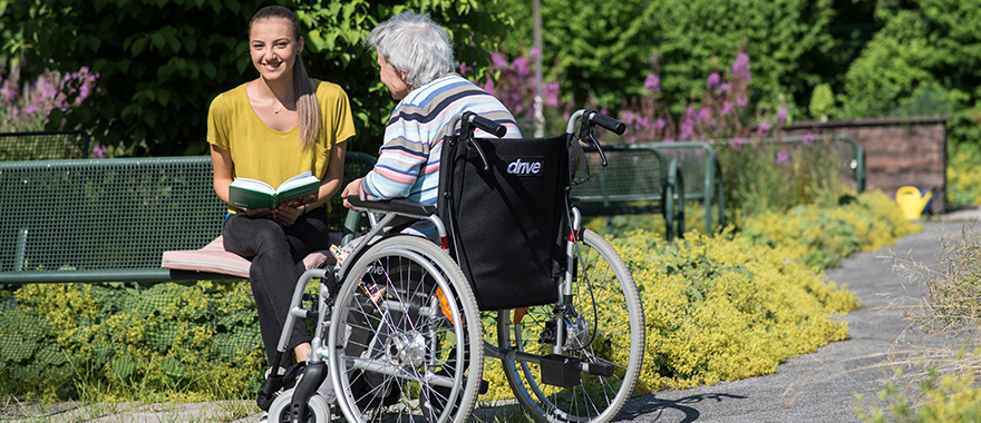 Eine junge Pflegerin sitzt mit einer gehbehinderten älteren Patientin im idyllischen Garten des Bezirksklinikums Obermain.